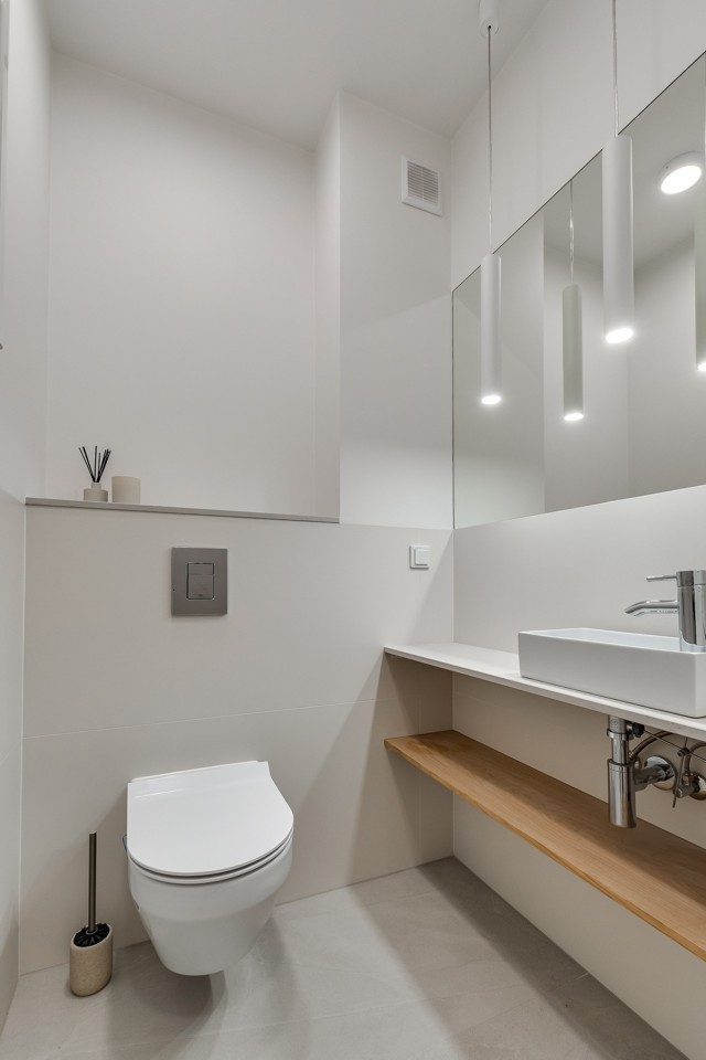 Minimalistyczna toaleta z bezową płytka na ścianie, mała nablatowa umywalka i drewnianą półką.